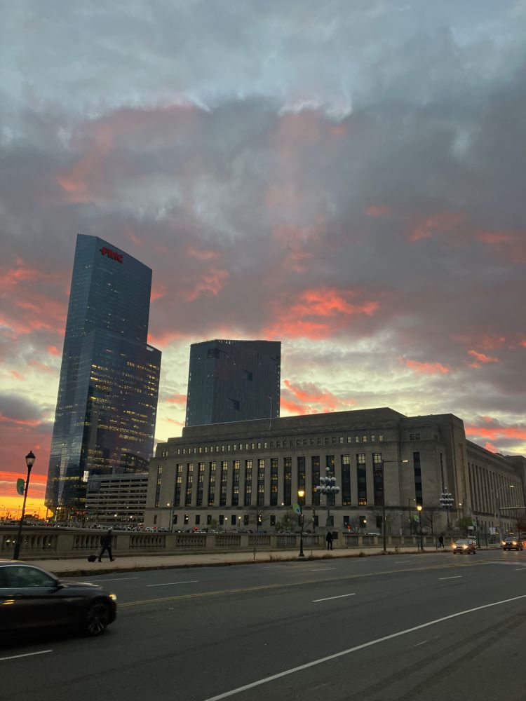 A photograph of sunset in Philadelphia on the evening of November 17, 2025, taken from the Market Street Bridge over the Schuylkill River. An arced line of a flock of birds is just about visible in the reddening sky. 