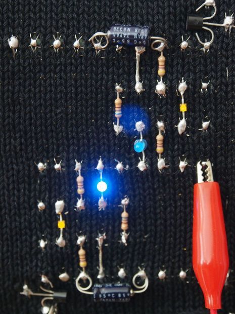 Close-up of a hand-knitted circuit board with an embedded LED lit up, showing conductive yarn forming the circuit paths on a small, patterned knitted fabric.