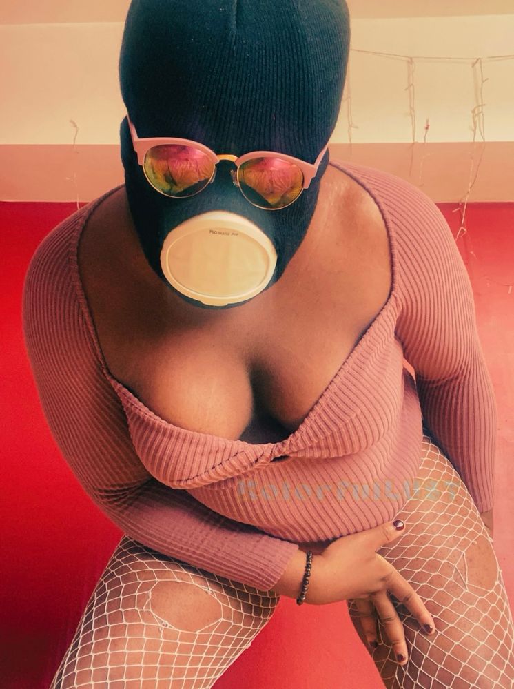 Image of Goddess Kolor standing in front of a red wall hunched over looking into the camera. Her face is obscured by a black ski mask, pink sunglasses, and a flo mask. She’s wearing a soft, cool toned, pink bodysuit, and her legs are seen with white fishnets sporting holes in them. Her fingernails are a deep red color.