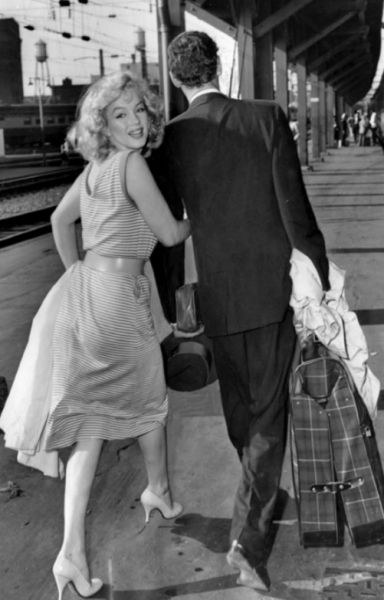 Black and white image of Marilyn Monroe in a light colored dress looking back over her shoulder while holding the arm of a man in a suit who is looking forward.