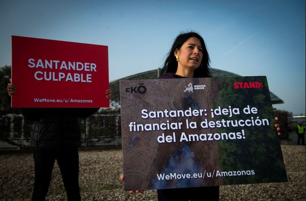 Stand.earth Senior Climate Finance Campaigner Martyna Dominiak holding a sign that reads “Santander: ¡deja de financiar la destrucción del Amazonas!

WeMove.eu/u/Amazonas” with Eko, WeMove Europe, and Stand.earth logos on it. while someone behind her holds a sign in front of their face that reads “SANTANDER CULPABLE WeMove.eu/u/Amazonas”