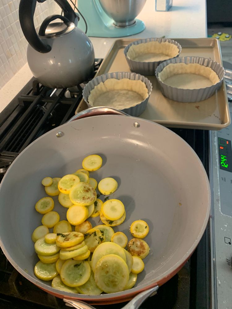 A sauté pan full of sliced squash next to a tray with prepared tart shells