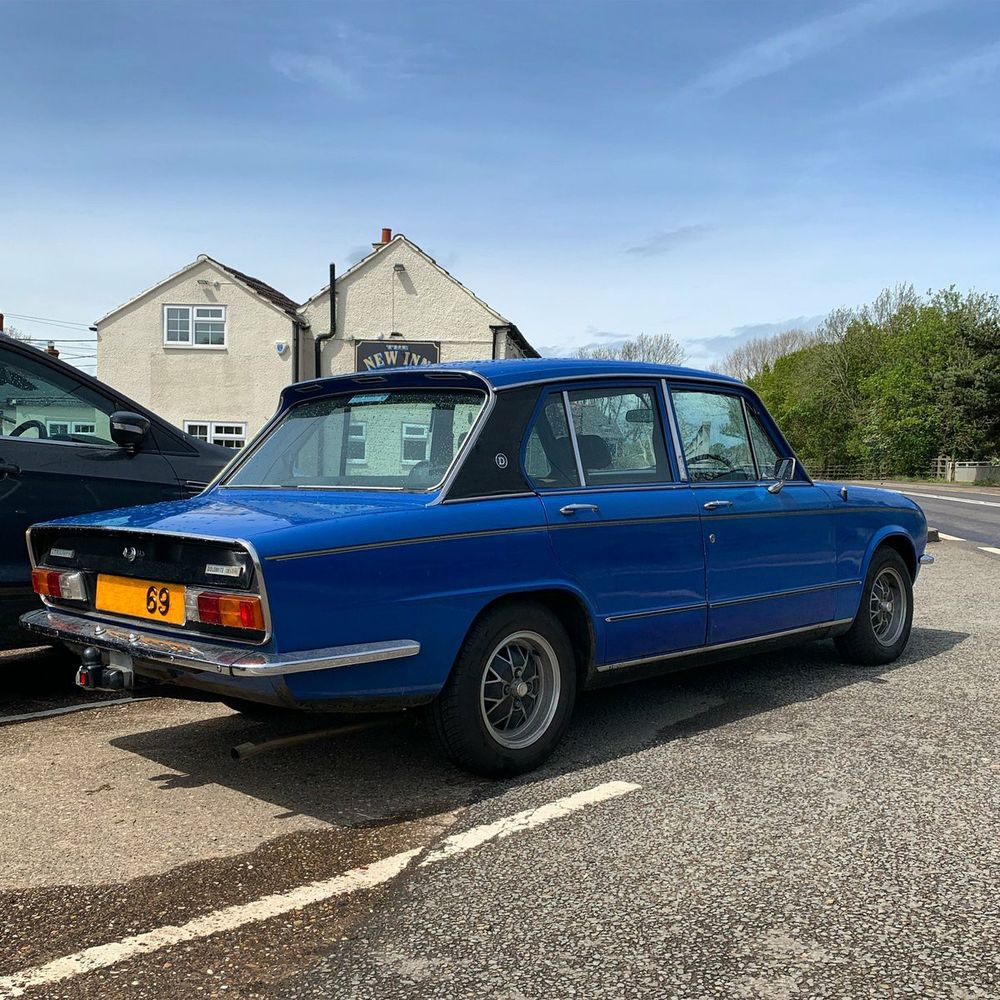 A photo of the back of a blue Triumph Dolomite with a black rear panel and C pillars in front of a pub