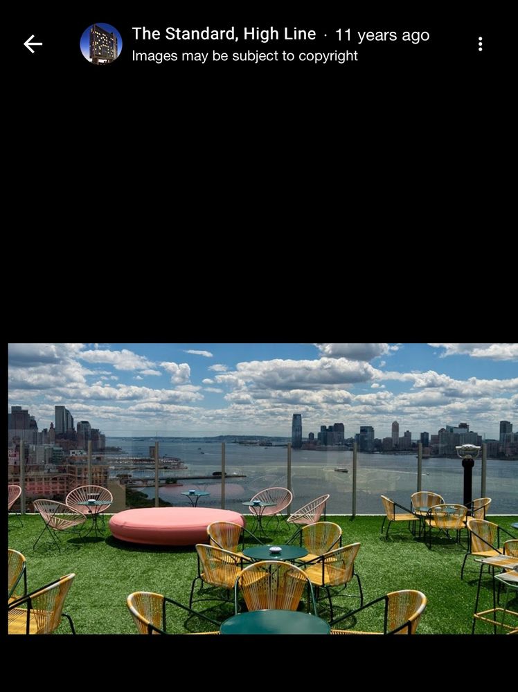 A balcony with several round tables with chairs. A large salmon colored resting area is in the rear of the photo. The balcony overlooks water and New York City skyscrapers in the background.