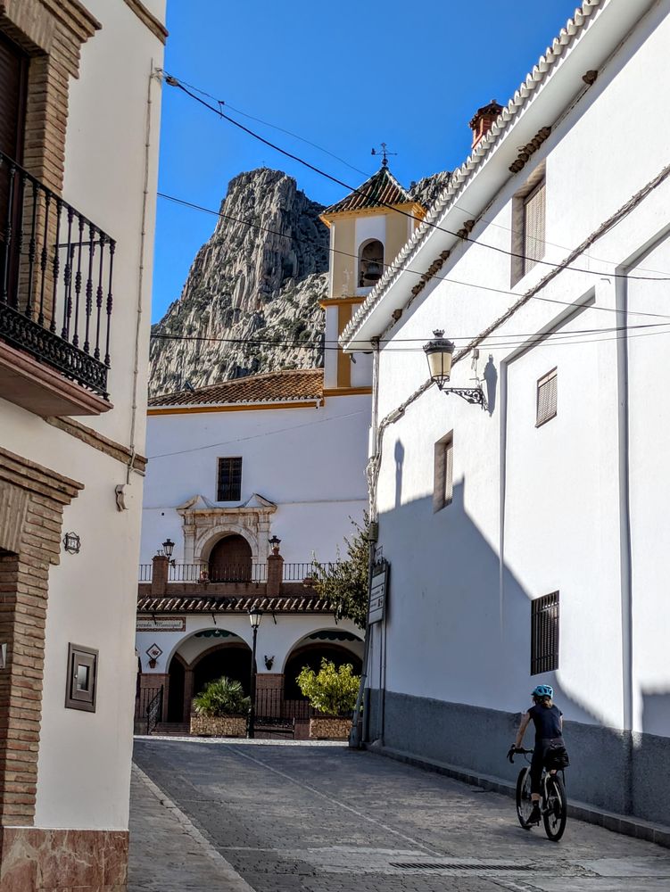 Woman riding a bike up a steep street with church and cliffs in the distance.