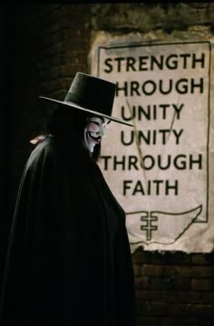 V from V for Vendetta, standing next to a sign that says “Strength Through Unity, Unity Through Faith”, ready to shake shit up. 