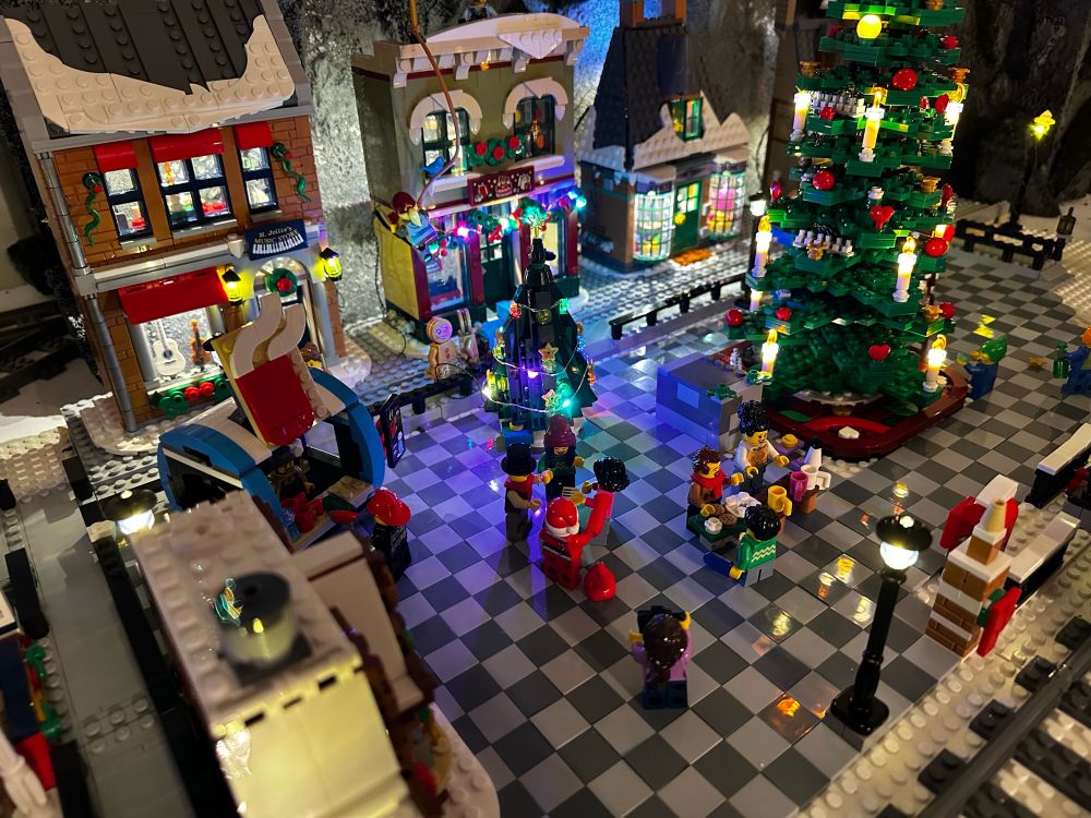 The city square of the village alternating light/dark gray tiles. Shops line a side with a giant Christmas tree in the center. 