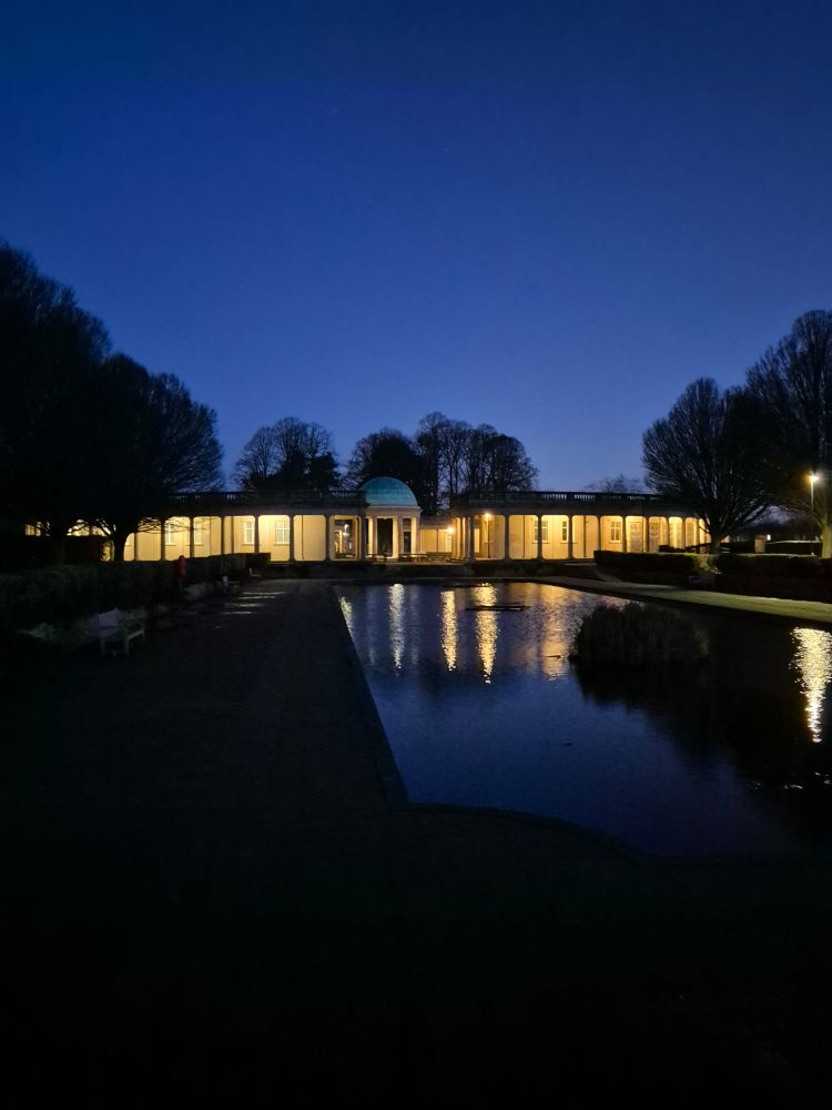 Eaton park. The view feom the bridge, over the pond towards the bandstand. Its still dark, the bandstand is lit up and the light reflects on the water. Its still and peaceful. 