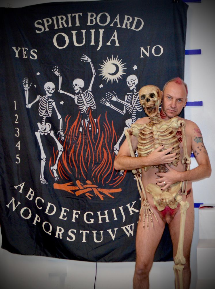 Male nude posing with Skelton and ouija board backdrop. 