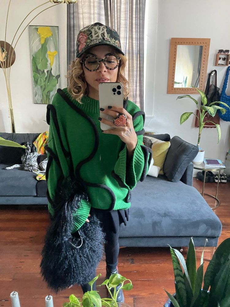Skye leaning in to take a mirror pic wearing an oversized green sweatshirt with 3D black squiggly lines all over it, round acetate glasses, a brocade looking Yankees hat, and a big copper filled heart ring. Holding a black furry lambskin purse