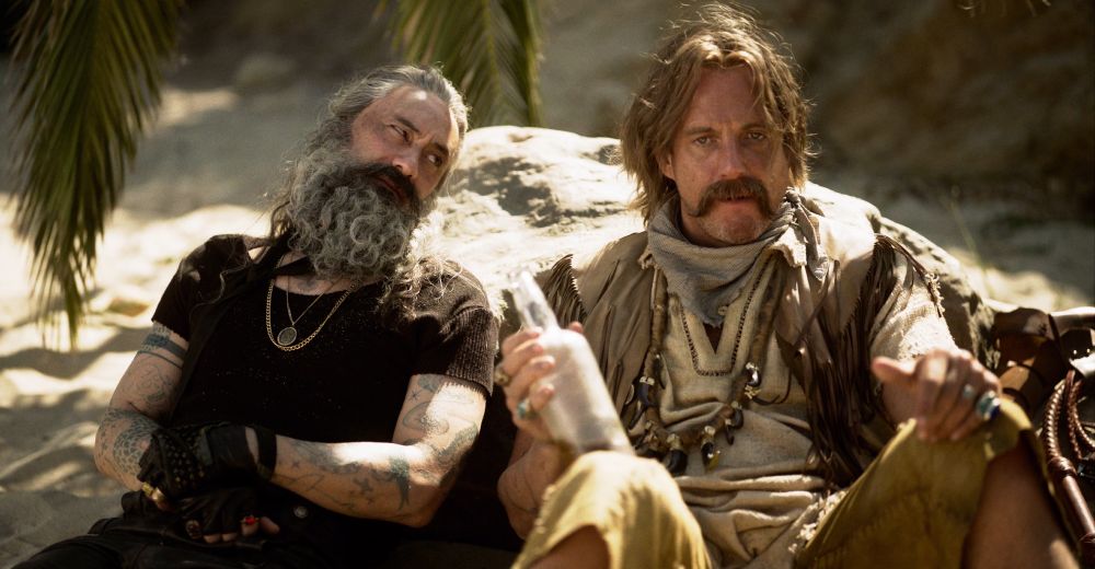 ed teach and calico jack sitting on the beach in the sand leaning against a rock, ed is glancing over with a bit of side-eye, calico jack is holding the rum (our flag means death s1e8)