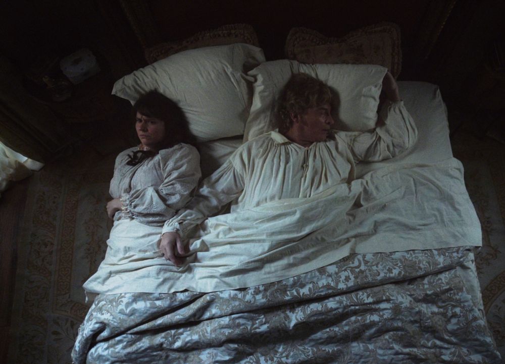 a shot looking down from above at stede bonnet in bed with mary, stede has stretched so far that he's nearly dead center in the bed with an arm outstretched almost pushing mary onto the floor, the look on her face is conspiracy to commit murder (our flag means death s1e10)
