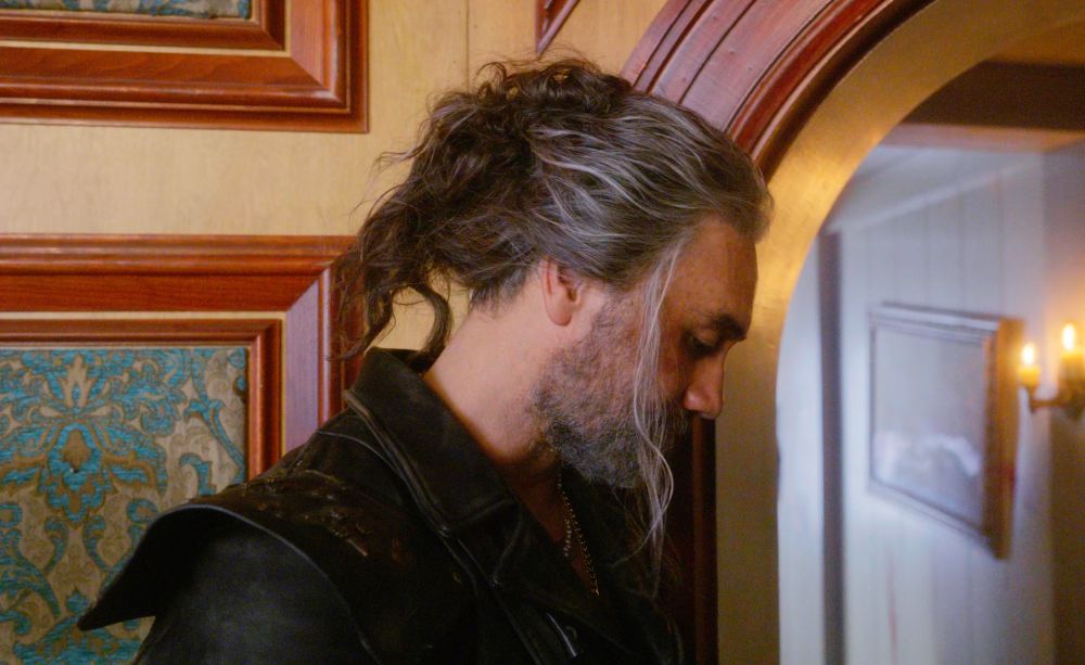 messy bun ed teach leaning against the wall, turning to look down giving a side profile of the bun and the slutty strands of hair (our flag means death s2e2)