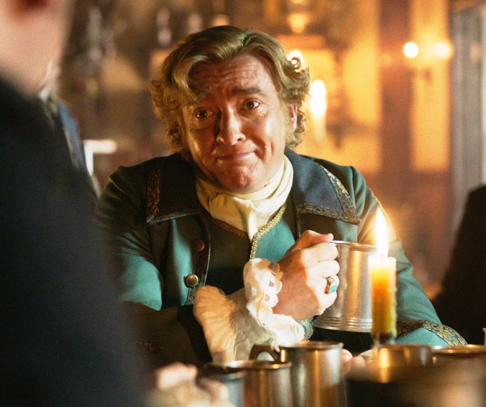 the gentleman pirate stede bonnet seated at a table holding a beer in a metal mug, smiling and bursting with joy as he thinks about how much he misses id (our flag means death s1e10)