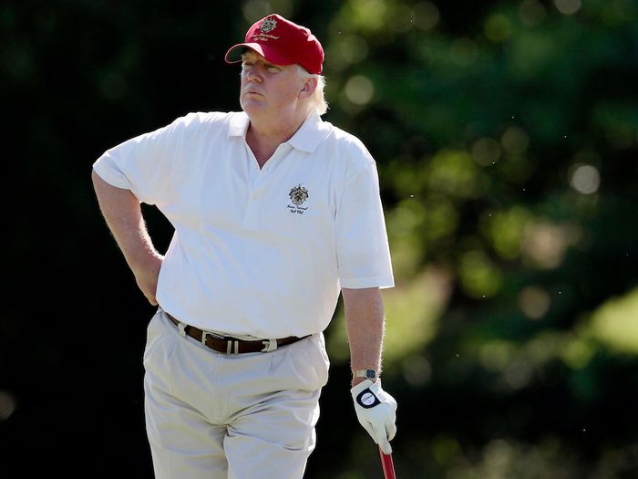 Photo of Trump wearing a white polo, khakis, and MAGA hat, leaning on his golf club with one hand, the other arm on his opposite hip. His physique is not what anyone would call “toned.”