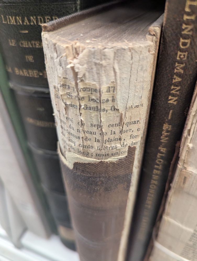 a damaged brown binder's volume. the outer spine has come off at the top, revealing a protective layer of reused paper with illegible French text printed on it, and the bare paper of the text block at the very top