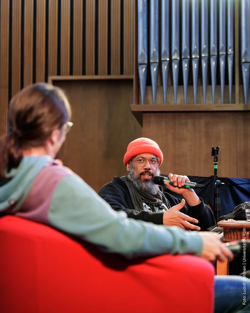 Eine Person mit einer roten Mütze spricht in ein Mikrofon und sitzt in einer Diskussionsrunde.