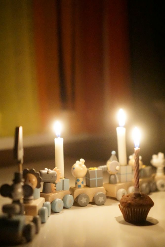 Ein Geburtstagszug aus Holz mit verschiedenen Tieren, farblich in grau und türkis gehalten. Auf dem ersten Wagen steckt die Zahl 4. Zwei Kerzen leuchten auf dem Zug. Vor dem Zug steht ein kleiner Schokoladenmuffin, ebenfalls mit einer Kerze drin. Im Hintergrund ist verschwommen ein Vorhang aus Kreppbändern in den Farben des Regenbogens zu sehen. 