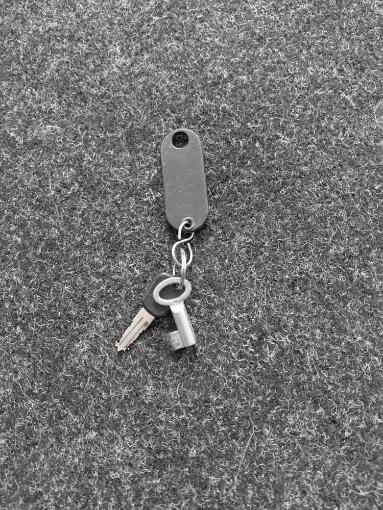 Fußboden, typischer Nadelfilz für Büro u d Schulen in Anthrazit. Darauf liegen zwei kleine Schlüssel, die an einem Schlüsselschild befestigt sind. Das Schild ist schwarz und liegt mit der Rückseite nach oben. 