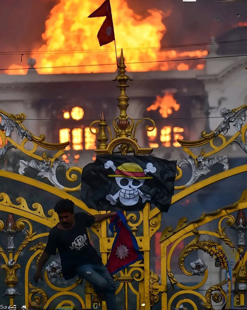 A nepalese protestor hanging the Straw Hat flag from One Piece on the gates of a government building as it's on fire behind him.