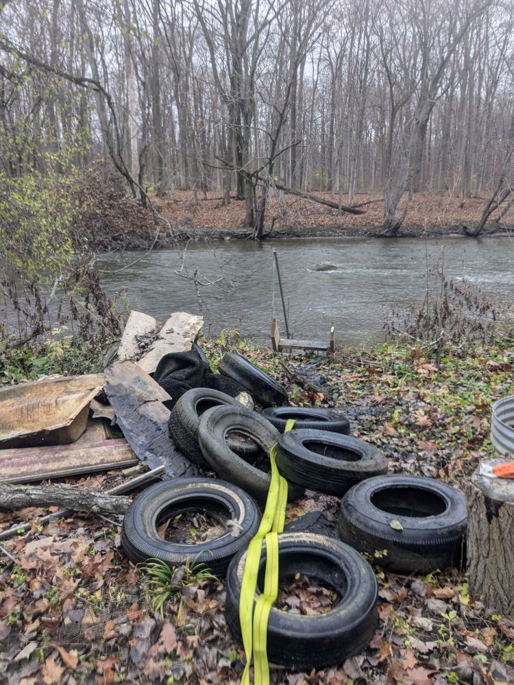 A pile of at least 10 tires and some other assorted trash next to a river. All of the tires and trash were in the river previously.