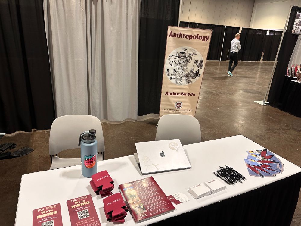 A picture of an exhibition booth at the AAA conference. There is a white tabletop with flyers, a pile of pens, stacks of notepads, and stacks of can koozies.
