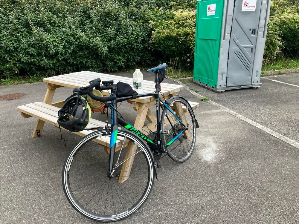 A black drip-handled bicycle leans against a wooden picnic table. A yellow helmet hangs from the handlebars. Behind these is a green hedge, and to the right is a green and grey site lavatory, like a tardis from a crapper dimension. 