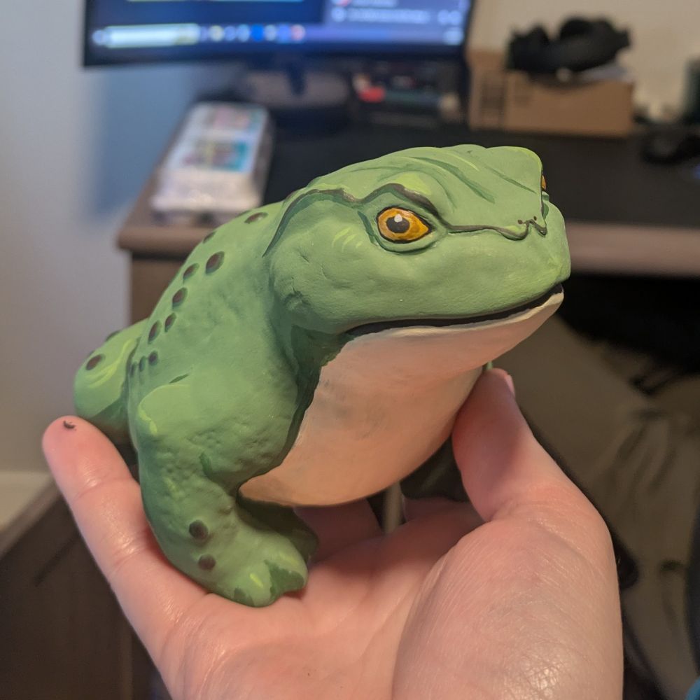 A clay frog painted with green skin, brown bumps, and a cream colored belly. Eyes are gold and looks a little angry.