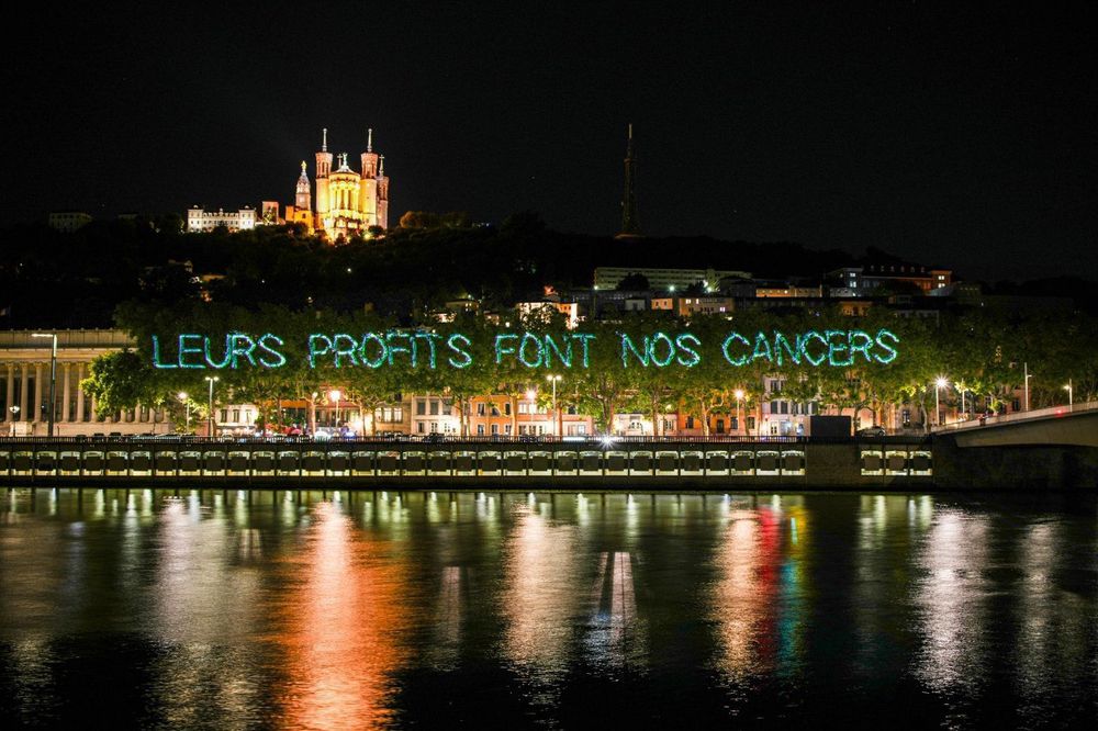 Photo prise de nuit
Au premier plan le rhone scintille 
Au second plan, En géant, la phrase « leurs profits font nos cancers » est projetée sur des arbres 
En arrière plan la basilique notre dame de la fourviere à Lyon 