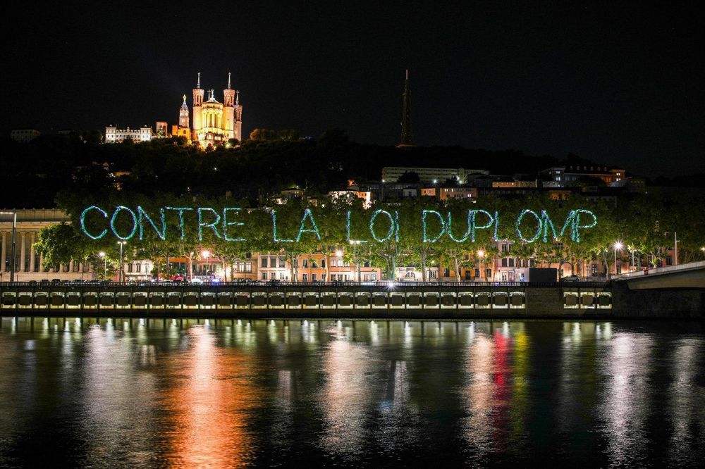 Photo prise de nuit
Au premier plan le rhone scintille 
Au second plan, En géant, la phrase « contre la loi duplomb » est projetée sur des arbres 
En arrière plan la basilique notre dame de la fourviere à Lyon 