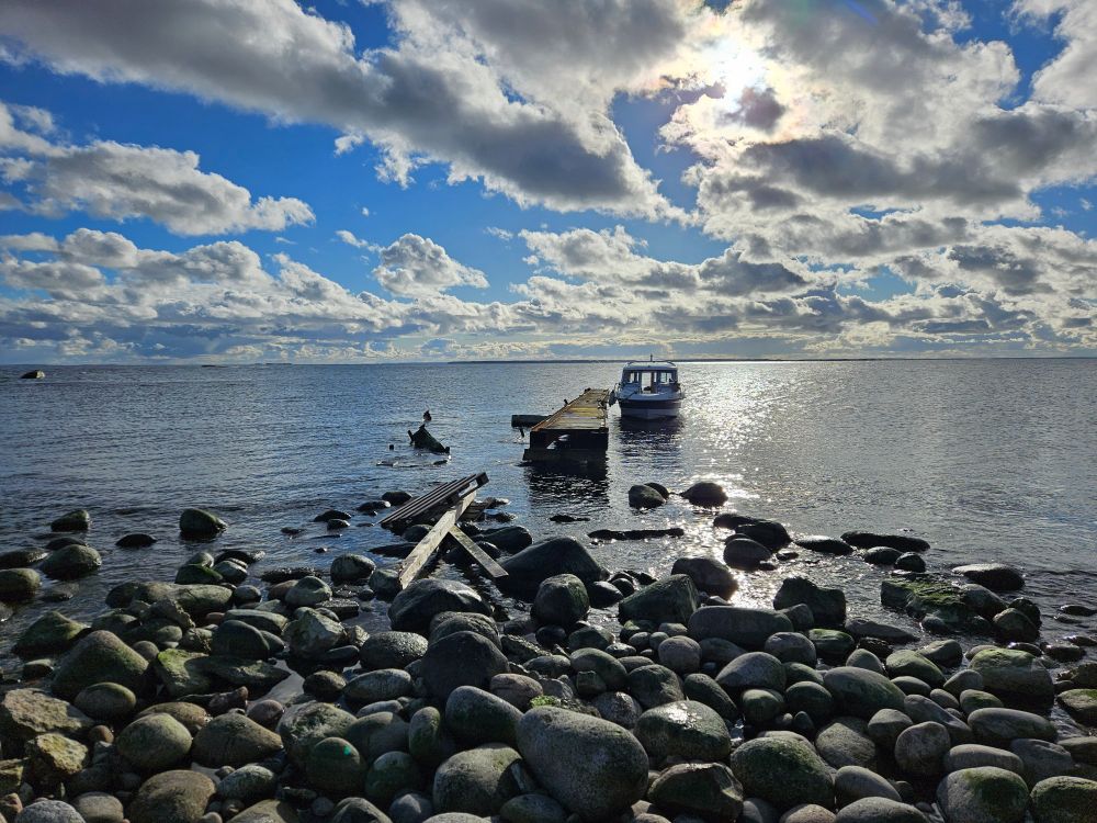 A beach of large (or medium-sized) round rocks, a jetty that does not come all the way to the beach, a small motor boat moored to the jetty, water, horizon, patchy blue sky and clouds with the sun shining behind the.