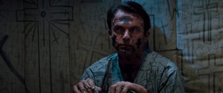 Still from In the Mouth of Madness of a man in a padded cell that’s dressed in a gown covered in black crosses 