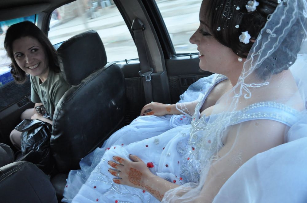Photo showing two women in a car, one wearing a white wedding dress with red accents and white veil