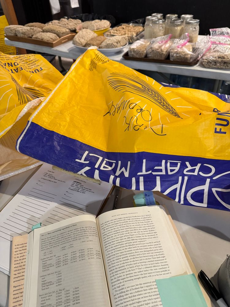 Photo showing an open book (In the Land of Ninkasi), a brewing plan for a golden and a dark beer, an empty bag of malted barley from Epiphany Craft Malt, and a table full of bappir cakes.