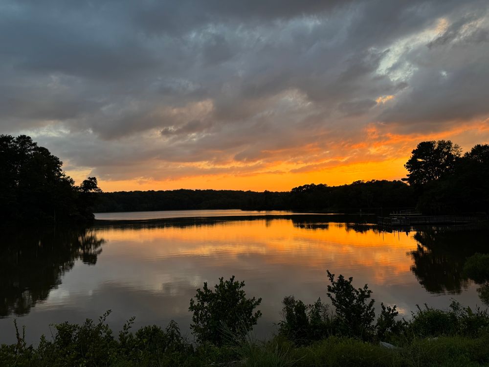 Photo showing a lake bordered by trees, just after sunset, with a cloudy sky (and mirrored lake surface) fading from bright orange to grayish purple