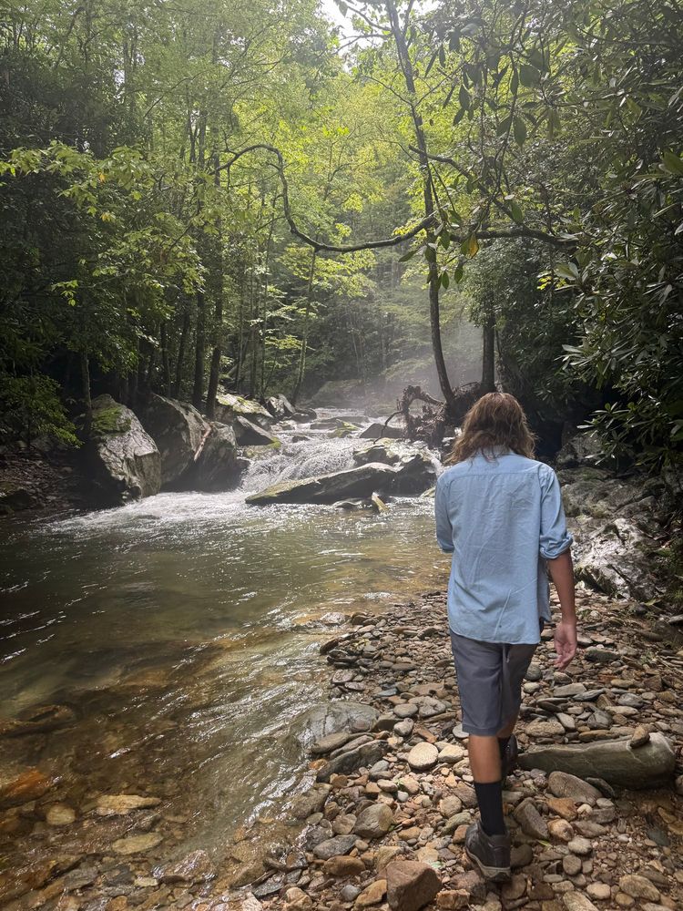Photo showing a man walking on pebbles alongside a mountain creek in dappled sunlight with large boulders, a small waterfall, green forest, and mist rising in the distance 
