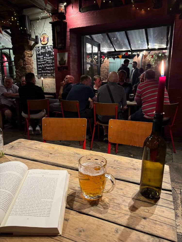 Photo in a pub showing a book, a pint of cask ale, and a candle in a wine bottle sitting on a wooden table