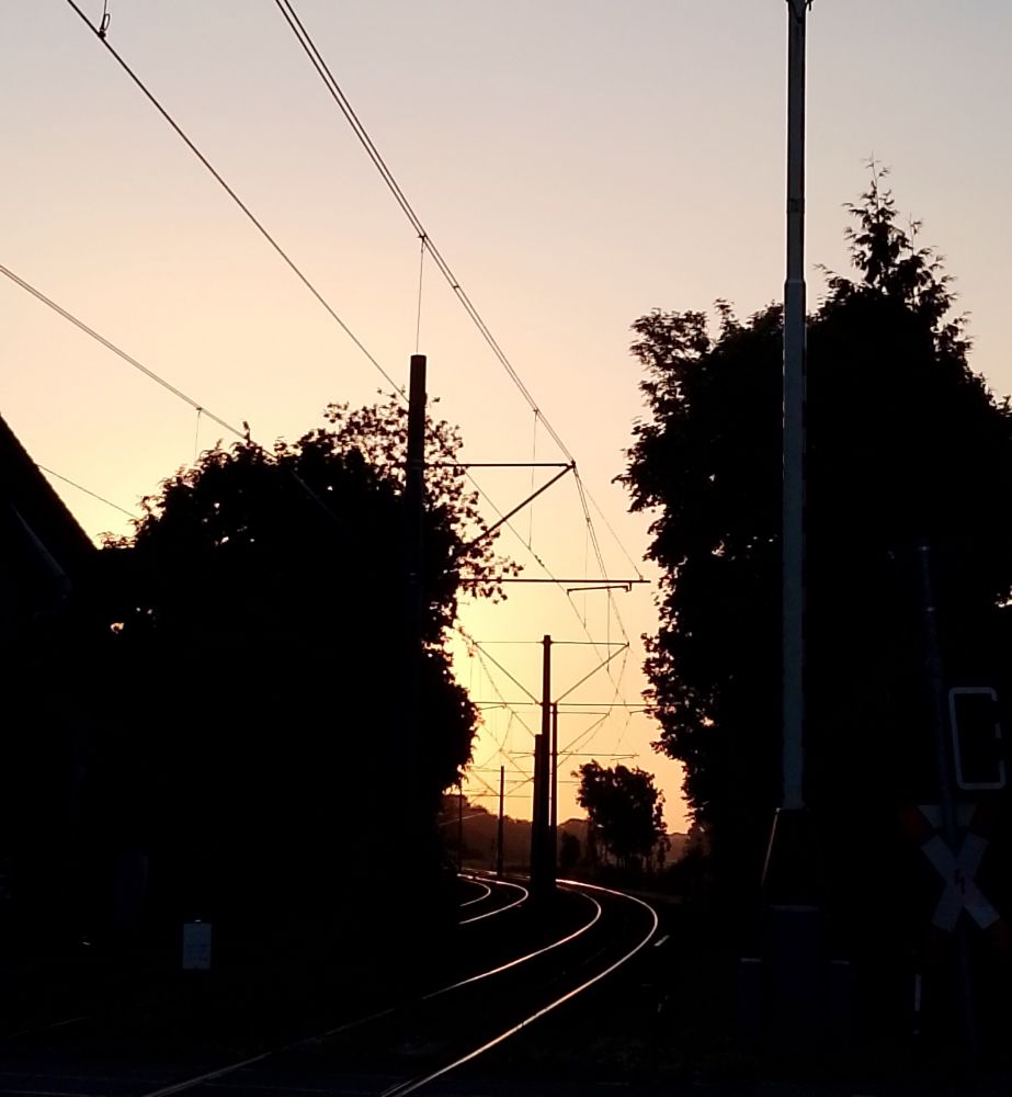 Foto wo die Sonne heute morgen aufgeht in schönen sommerlichen Farben, wie in Orange, Gelb, etwas lila. In der Mitte des Bildes sieht man Tramgleise. Foto wurde in der Nähe von Düsseldorf gemacht , am Bahnübergang. 