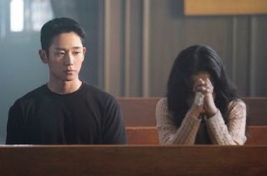 sitting man in a black tshirt staring down at a woman who is also sitting clasping her hands together in prayer 