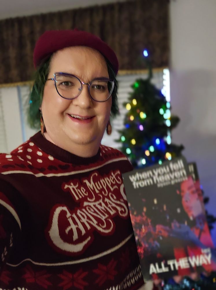 Selfie. White woman. Burgundy beret.  Green hair with a little tinsel in it.  Muppet Christmas Carol sweater and earrings.  I'm in front of a Christmas tree and I'm holding 2 copies of When You Fell From Heaven book 2. 