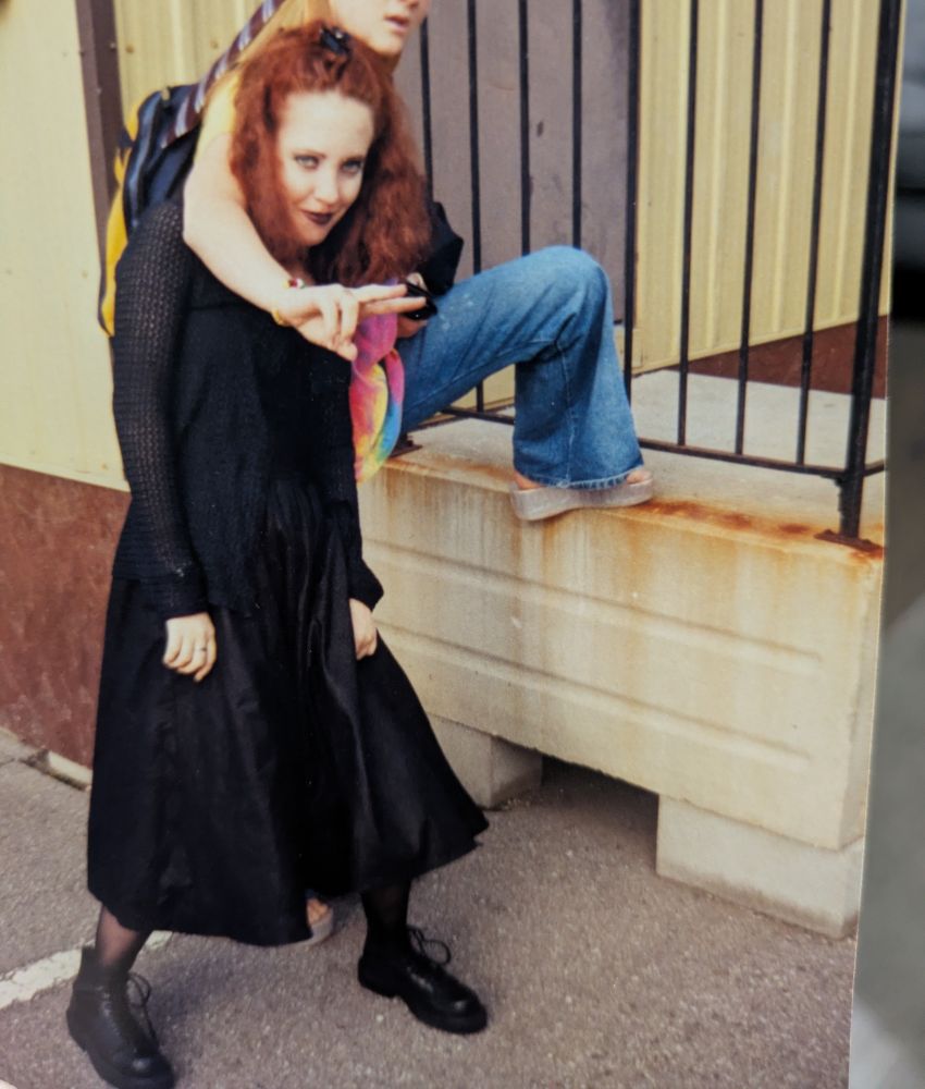 A teenager with bright red frizzy hair stands in a black dress, black boots, and black lipstick while someone dressed as a hippy stands behind them giving a peace sign