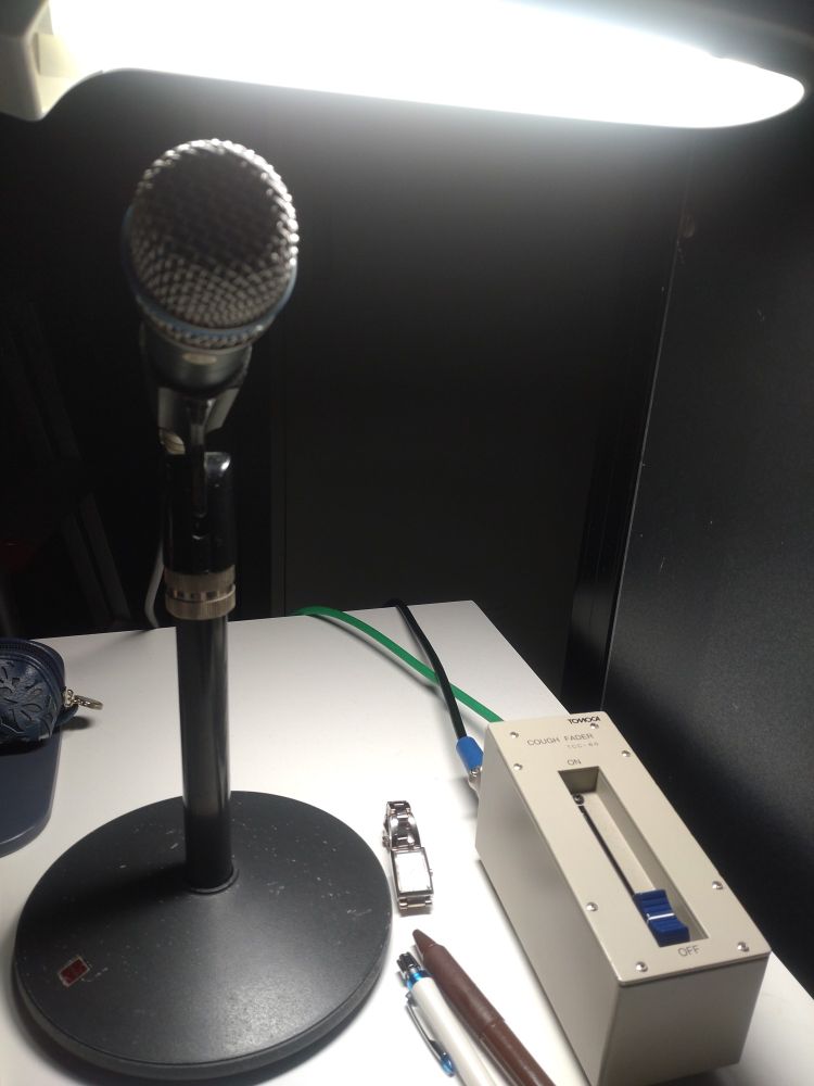 小さなデスクの上にマイクとライトとペンと時計。見えないけど手前に原稿がある。某所舞台袖。