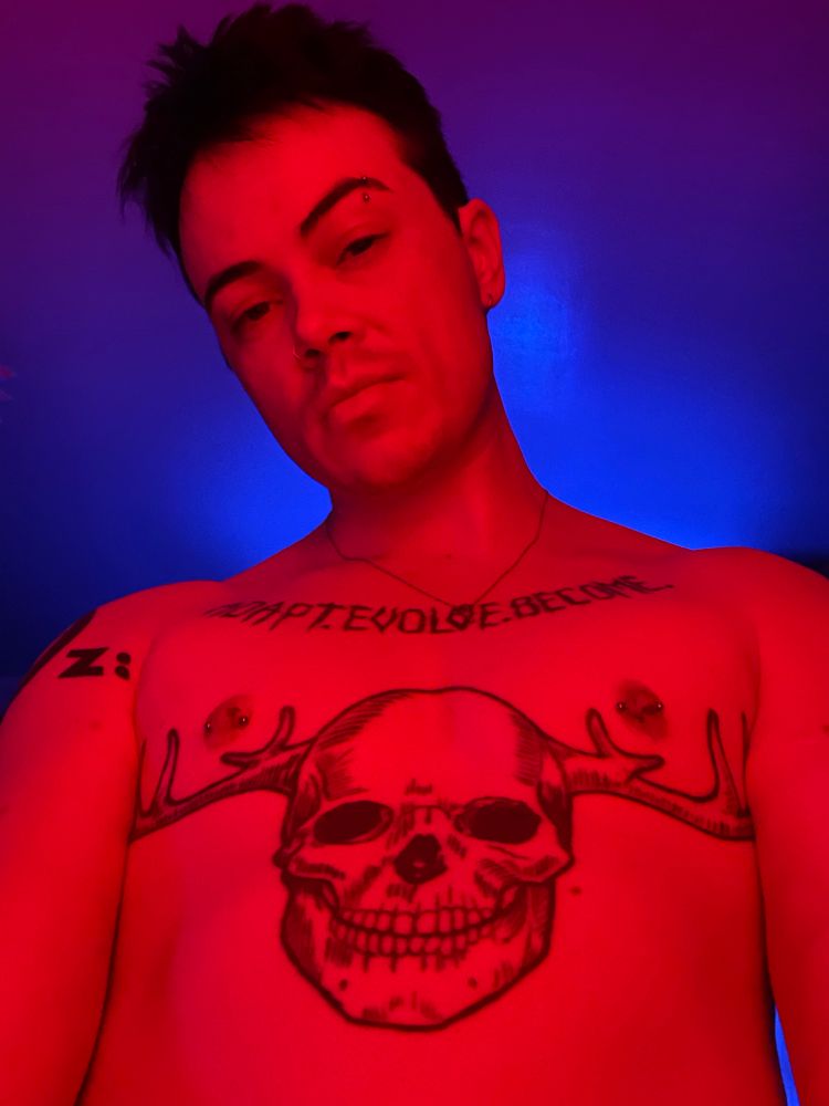 Damian shirtless in his bedroom lit by intense red light while the room behind him is lit up blue 