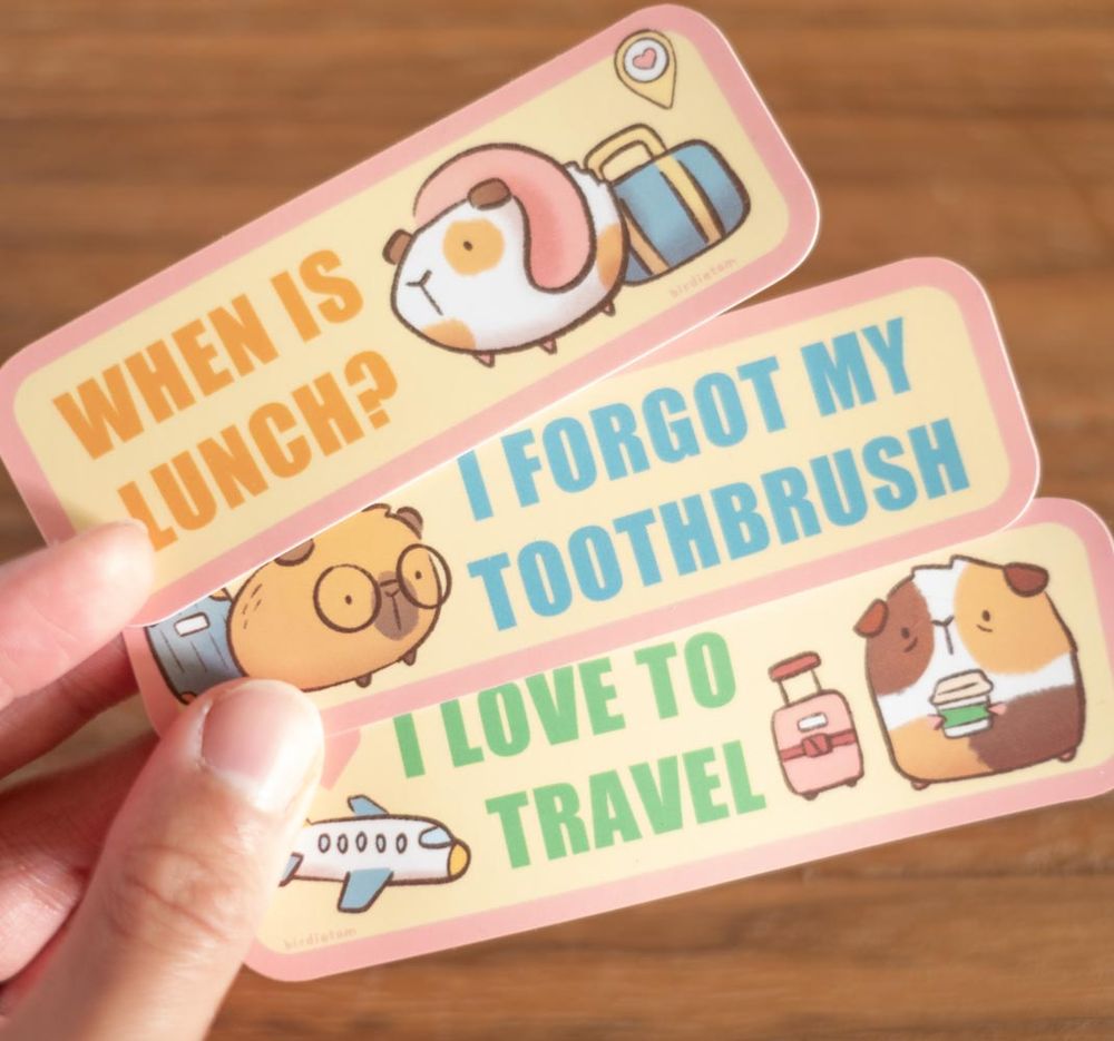 A photo of a hand holding 3 stickers with images of guinea pigs traveling. The text on them read "When is Lunch," "I forgot my Toothbrush," and "I love to travel."