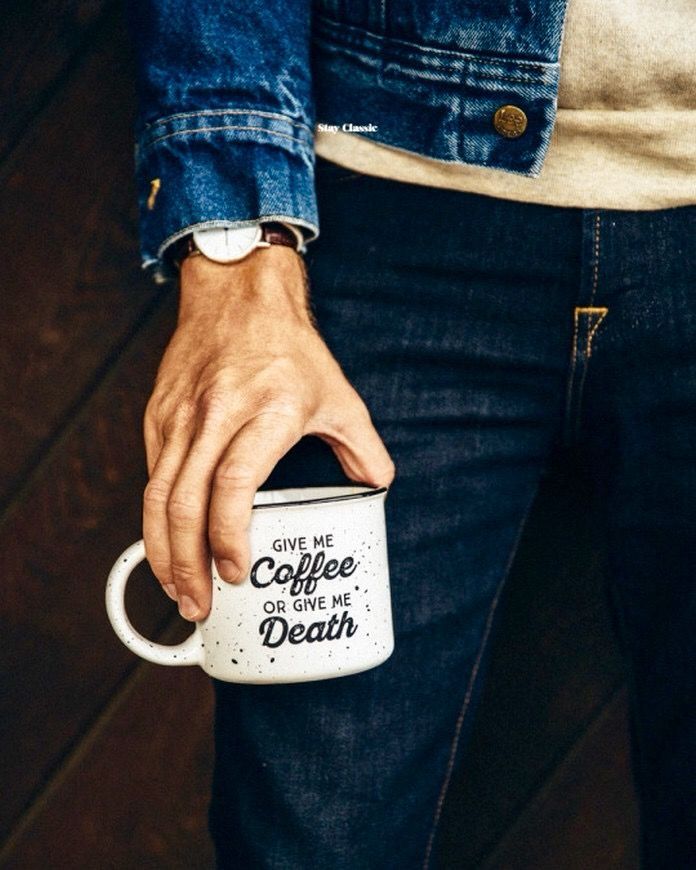 Een hand van een persoon die een mok vasthoudt. De mok is wit met zwarte spikkels en heeft de volgende tekst in zwarte letters erop staan: "GIVE ME Coffee OR GIVE ME Death". De persoon draagt een blauw jeansjack en een donkere jeansbroek. Aan de pols zit een horloge met een bruine leren band. De achtergrond is donkerbruin en houten planken zijn zichtbaar.