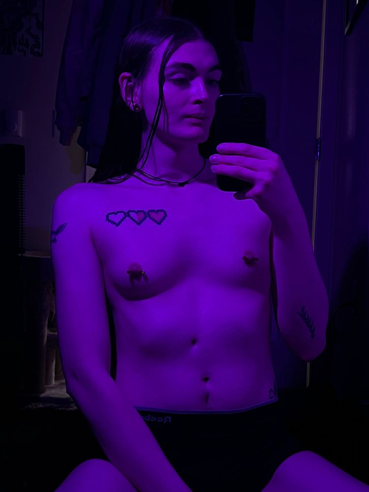 A topless selfie of a woman sitting on her floor in front of her mirror, the room is lit by purple lighting 