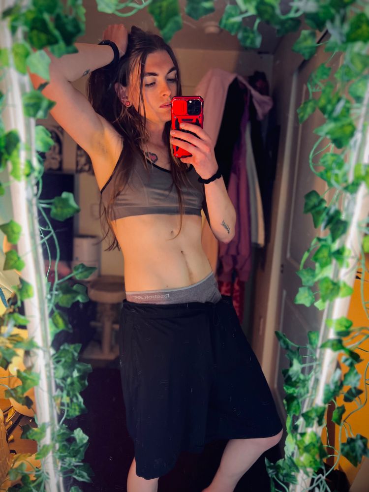A woman with long brown hair, Zai, takes a selfie in a mirror decorated with green leafy vines. She is wearing a gray sports bra, black shorts, and lavender underwear peeking out above the waistband of her shorts. She holds her red phone up to take the photo while one hand rests on her head, her arm bent at the elbow. She has tattoos on her arms and a small tattoo near her collarbone.