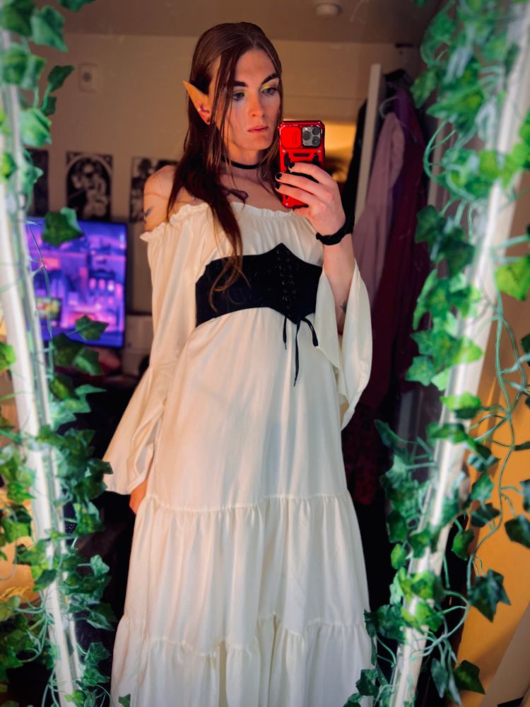A full-length mirror selfie of a woman dressed as an elf. She has long, light brown hair, and is wearing prosthetic pointed elf ears. She is dressed in a white peasant-style dress with a tiered skirt and billowy sleeves, cinched at the waist with a black, corset-style belt. She is holding a red smartphone to take the photo. Her choker necklace and a tattoo on her left shoulder are visible. The mirror is framed with artificial green ivy, and in the background, a TV screen and a clothes rack are visible.