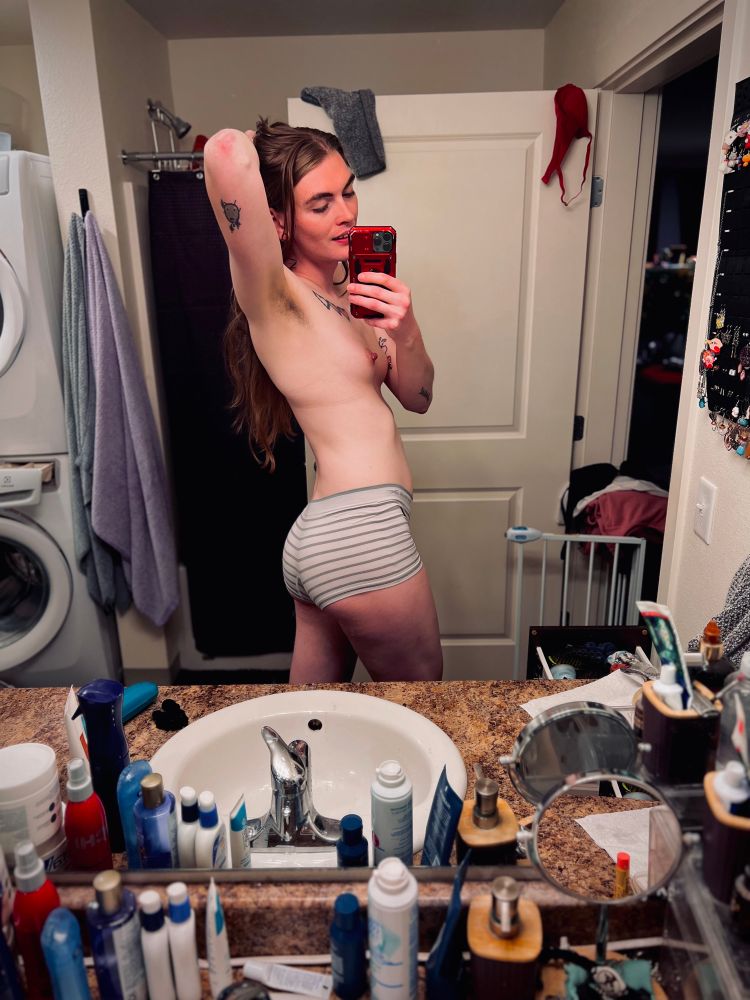 A woman with long brown hair, Zai, takes a selfie in a bathroom mirror. She is holding her hair up with one hand while holding a red phone with the other. She is wearing light gray and white striped underwear, and her torso is bare. Her tattoos are visible on her arms and side. A full counter of hygiene products sits in the foreground, and behind her is a washing machine and various clothes hanging.