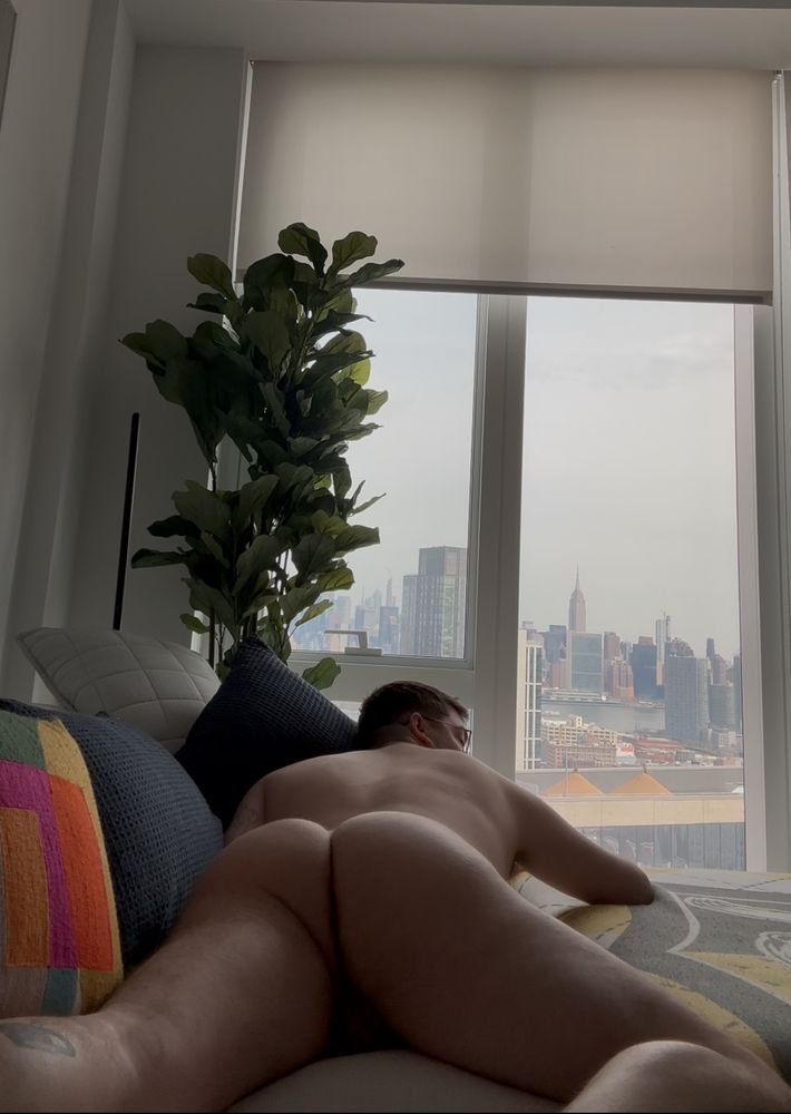 facing a window that looks over a city skyline, a man lays on his stomach naked, with one leg bent up displaying his butt and back 